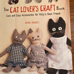 May include: The cover of "The Cat Lover's Craft Book" features three handmade cat dolls wearing dresses. The book's title is in red, with the subtitle "Cute and Easy Accessories for Kitty's Best Friend." The cover also shows cat-themed accessories.