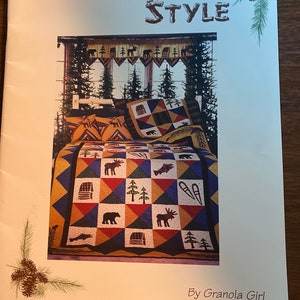 Op de afbeelding: Een quiltpatroonboek met de titel "Northwoods Style" van Granola Girl. De omslag toont een kleurrijke quilt met een bosmotief, waaronder elanden, beren, bomen en hutten.