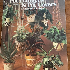 May include: A book cover with the title "Crocheted Pot Hangers & Pot Covers" by Barbara Johnston. The cover features a variety of crocheted plant hangers and pot covers in different colours and styles.