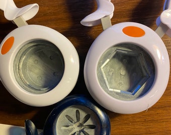May include: Three different paper punch tools. One is white with an orange handle and a round punch. Another is white with an orange handle and a hexagonal punch. The last one is black with a blue handle and a flower-shaped punch.