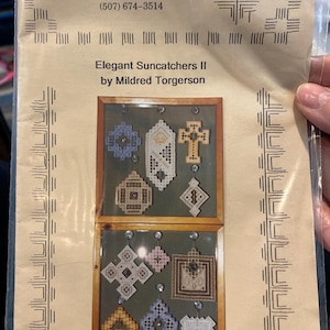 Op de afbeelding: Een knutselpatroon getiteld "Elegant Suncatchers II" door Mildred Torgerson. Het patroon toont verschillende geometrische ontwerpen in een vitrine. De tekst "Hardanger-Millie's Way" en contactgegevens zijn ook zichtbaar.