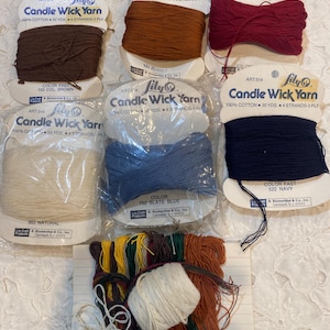 May include: A selection of Lily Candle Wick Yarn in shades like brown, russet, navy, and slate blue. The image displays several spools and bundles of yarn, ideal for crafting and textile projects. The packaging indicates 100% cotton, with lengths of 27-46 metres.