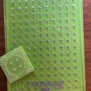 Op de afbeelding: Een rechthoekige limoengroene Cuttlebug-embossingplaat met een raster van kleine cirkelvormige inkepingen en een paars logo met een pijl die naar beneden wijst. Een vierkante limoengroene embossingmap met een decoratief cirkelvormig patroon ligt erbovenop.