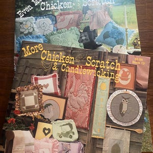 May include: Two books are visible, titled "Even More Chicken Scratch" and "More Chicken Scratch & Candlewicking". The covers display cross-stitch and embroidery projects, such as pillows and framed designs. The books are presented on a wooden surface.