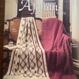 Op de afbeelding: Een boekomslag met de titel "Heirloom Afghans" met twee dekens op een stoel. Eén is crèmekleurig met bloempatronen en franjes, de andere is effen bordeauxrood met een gestructureerd ontwerp. De tekst op de omslag luidt "In Knit, Crochet and Afghan Stitch by Marion Graham."