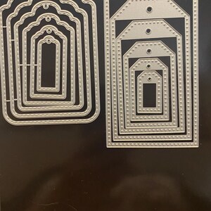 May include: A set of silver metal cutting dies in various sizes and shapes. One set features a scalloped edge design, while the other set has a tag shape with a hole at the top. These dies are likely used for paper crafting and scrapbooking projects.