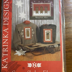 May include: A package featuring a winter-themed design with the text "Snowflake Flyer." The design includes a sweatshirt, a basket, and a framed image, all with a sledding theme. The package is sealed in clear plastic.