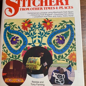 May include: A vintage book cover titled "Here and Now Stitchery From Other Times & Places." The cover features colorful embroidery designs, including floral patterns and a central image of a person with a backpack. The book is by Robbie and Tony Fanning.