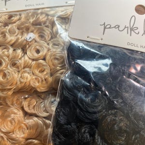 Puede incluir: Se muestran dos paquetes de pelo de muñeca. Uno contiene cabello rubio claro y rizado, y el otro cabello negro y rizado. Los paquetes tienen una etiqueta blanca con las palabras "park lane" y "DOLL HAIR" impresas.