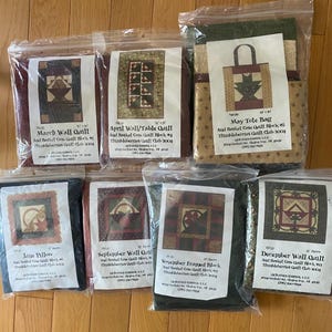 May include: A collection of packaged quilt blocks and tote bags, each labeled with a month and quilt type. The packages feature various quilt designs with floral and geometric patterns in shades of red, green, and brown. The text on the packages includes the names of the quilt blocks.