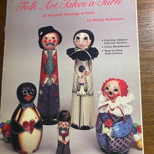 May include: A book cover titled "Folk Art Takes a Turn" featuring painted wooden figures of clowns and a penguin. The figures are colorful and detailed, with various patterns and designs. The book is part of the Collectors Series.
