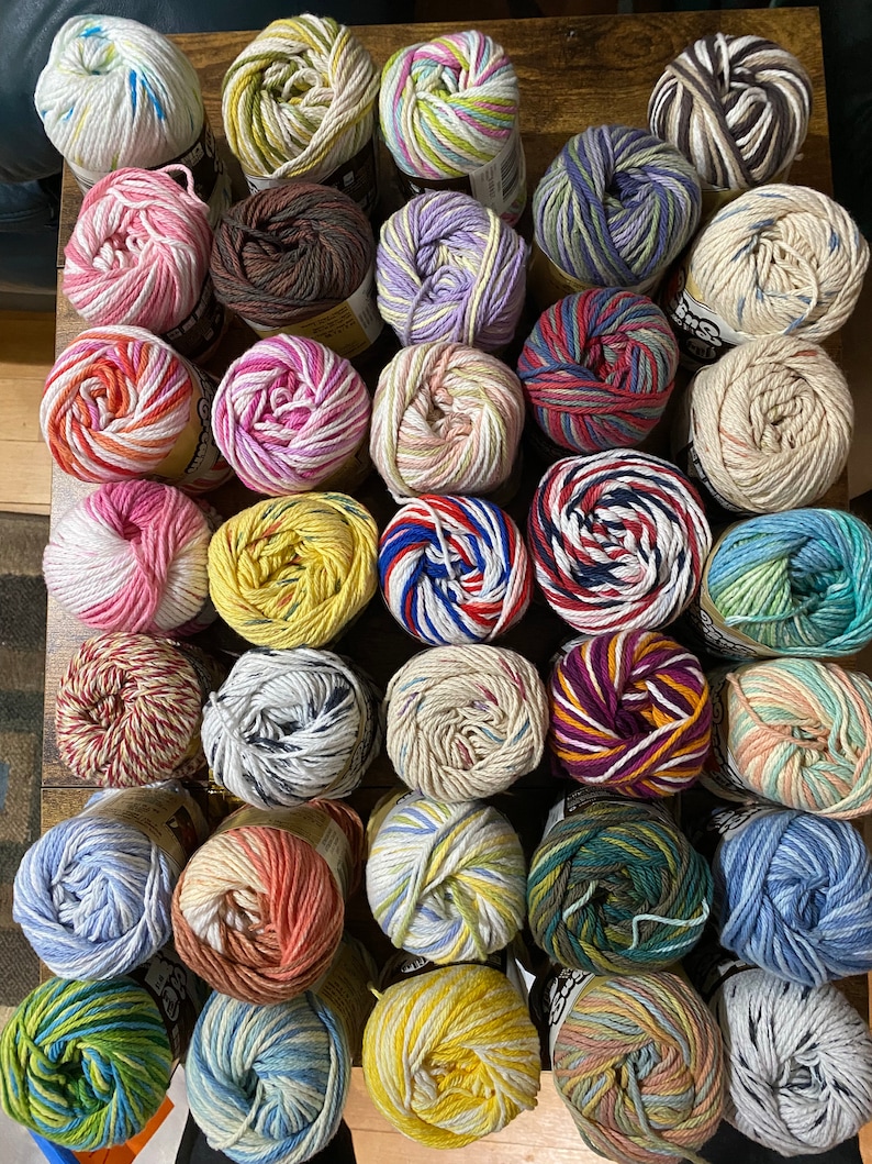 May include: A collection of 36 colorful yarn balls, each with a different pattern and color combination. The yarn balls are arranged in rows on a wooden surface.