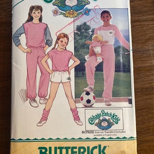 Op de afbeelding: Een vintage Butterick naaipatroon voor Cabbage Patch Kids outfits. De omslag toont illustraties van kinderen in roze en witte outfits, met een voetbal en een Cabbage Patch Kids pop. Het patroon bevat strijkapplicaties.