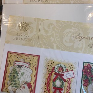 Może przedstawiać: Zestawy kart świątecznych warstwowych Anna Griffin. Karty mają skomplikowane złote filigranowe ramki z świątecznymi wzorami. Jedna karta przedstawia Świętego Mikołaja i choinkę, inna wieniec z czerwonymi kokardami, a trzecia poinsecję.