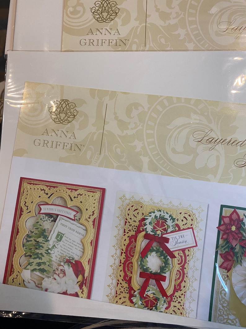 Może przedstawiać: Zestawy kart świątecznych warstwowych Anna Griffin. Karty mają skomplikowane złote filigranowe ramki z świątecznymi wzorami. Jedna karta przedstawia Świętego Mikołaja i choinkę, inna wieniec z czerwonymi kokardami, a trzecia poinsecję.