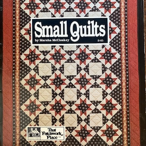 Puede incluir: Portada del libro "Small Quilts" de Marsha McCloskey. La portada presenta un diseño de edredón con estrellas y cuadrados rojos, negros y crema. El libro es publicado por That Patchwork Place.
