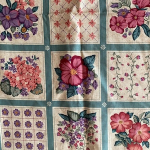 Op de afbeelding: Bloemenstof met een herhalend patroon van vierkanten. Elk vierkant bevat een ander bloemmotief in roze, paarse en groene tinten, tegen een crèmekleurige achtergrond en omlijst door een turquoise rand. De stof heeft een vintage uitstraling.