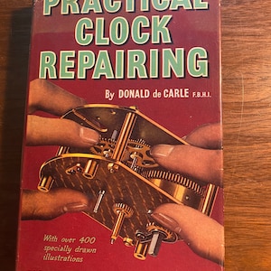 Op de afbeelding: Een vintage boekomslag met de titel "PRACTICAL CLOCK REPAIRING" door Donald de Carle F.B.H.I. De omslag toont een close-up afbeelding van een uurwerk met handen die de tandwielen vasthouden. Het boek bevat meer dan 400 illustraties en is uitgegeven door N.A.G. PRESS LTD. LONDON.