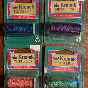 Könnte beinhalten: Vier Spulen Kreinik Metallics 1/8 Zoll Band. Die Spulen sind in Klarplastik mit grünen und weißen Etiketten verpackt. Die Bandfarben sind blau, lila, rot und grün.