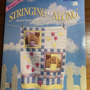 May include: A book cover titled "Stringing Along" with Vanessa-Ann. The cover features a quilt design with hearts and stars against a blue sky background. The book is part of the Designer Series by Trice Boerens.