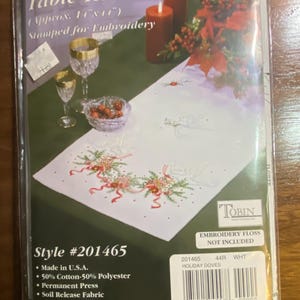 Op de afbeelding: Een witte Holiday Doves tafelloper, gestempeld voor borduurwerk, met een ontwerp van duiven, rode linten en groen gebladerte. De verpakking bevat de tekst "Approx. 36cm x 104cm" en "Made in U.S.A."