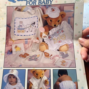 May include: A booklet with 47 cross-stitch patterns for baby gifts. The cover features a teddy bear wearing a hat and holding a rattle. The booklet includes designs for a diaper bag, a bib, a blanket, and a hat. The text on the cover reads "Quick Gifts for Baby".
