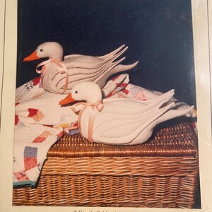 May include: Two white fabric geese with pink ribbon accents are sitting on a wicker basket. The basket is covered with a patchwork quilt. The image is from a vintage craft pattern.