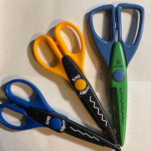 May include: Three pairs of decorative scissors with different coloured handles and cutting edges. The scissors are blue, yellow and green. The blades are black and have a zig-zag pattern. The scissors are labelled "Kraft Edgers".