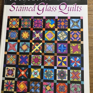 Op de afbeelding: Een softcover boek getiteld Foundation Pieced Stained Glass Quilts door Liz Schwartz & Stephen Seifert. De kaft toont een raster van kleurrijke, geometrische quiltblokken gescheiden door zwarte stroken, lijkend op glas-in-lood.