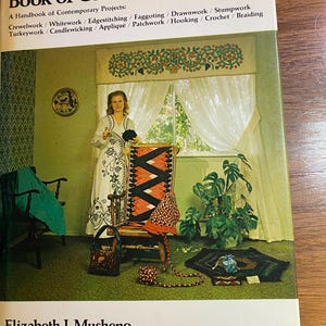 May include: The cover of "Book of Colonial Needlework" features a woman in a white dress, standing near a chair with a patterned seat. The book's title is in large letters, with a list of needlework techniques below. The author's name is at the bottom.