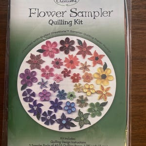 Può includere: Un kit di quilling Flower Sampler in una confezione sigillata. Il kit include istruzioni e carta per creare fiori di carta. L'immagine mostra una varietà di fiori di carta colorati in una disposizione circolare. Il testo sulla confezione recita "Quilled Creations".