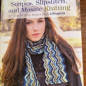 May include: A book cover titled "First Time Stripes, Slipstitch, and Mosaic Knitting" featuring a woman wearing a blue, white, and brown knitted scarf. The book cover has a light background with the text "Step-by-Step Basics Plus 3 Projects".