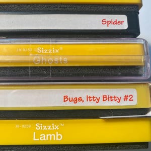 May include: Four yellow plastic storage containers with black lids. The containers are labeled with the text "Sizzix" and the names "Bird", "Ghosts", "Lamb", and "Bugs, Itty Bitty #2". The containers are stacked on top of each other.