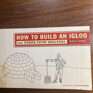 May include: A book titled "HOW TO BUILD AN IGLOO and OTHER SNOW SHELTERS" with illustrations. The cover features a drawing of an igloo, a person with a shovel, and the author's name. The book is on a wooden surface.