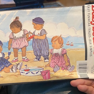 Könnte beinhalten: Ein Schnittmuster zum Nähen von Kinderkleidung. Das Muster ist für ein Set aus drei Outfits, jedes mit einer anderen Farbe und einem anderen Muster. Die Outfits sind alle für den Sommer gedacht und haben einen Matrosenkragen. Das Muster ist von Busybodies by Butterick und ist in den Größen NB-S-M.