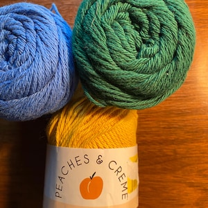 May include: Three balls of yarn in blue, green, and yellow. The yellow ball has a label reading "Peaches & Creme" with a peach graphic and "Original" text. The yarn is tightly wound, suitable for knitting or crochet projects.