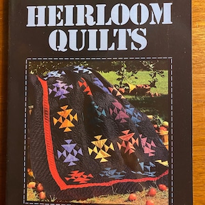 Puede incluir: Portada de libro con el título "HEIRLOOM QUILTS" en letras blancas grandes sobre fondo negro. La portada presenta un edredón colorido con patrones geométricos y el texto "A LEISURE ARTS PUBLICATION".