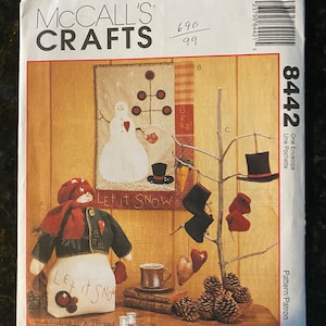 Op de afbeelding: Een vintage McCall's Crafts naai-patroon voor een sneeuwman-versiering. Het patroon bevat instructies voor het maken van een sneeuwman met een rode muts, een rode sjaal en een groen jasje. Het patroon bevat ook instructies voor het maken van een kleine boom met ornamenten, een kopje warme chocolademelk en een stapel boeken. Het patroon is getiteld "Let It Snow" en is genummerd 8442.