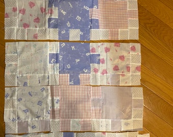May include: A patchwork quilt in progress, featuring squares of various fabrics. The quilt top includes sections of light blue, pink, and white fabrics with floral, letter, and polka dot patterns. White strips and squares are laid out below the quilt top.