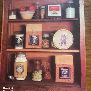 May include: A book cover titled "COUNTRY CUPBOARD" with a wooden cabinet design. The cabinet shelves display various vintage food items, including Campbell's Tomato Soup, jars of spices, and a box of Morton Plain Salt. The text "Book 8" is visible.