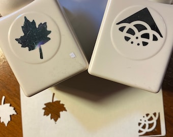 May include: Two white paper punch tools with black designs. The left punch has a maple leaf design. The right punch has a circular design with a centre dot and a triangle shape at the top.