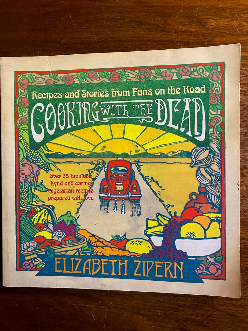 Puede incluir: Una portada de libro de cocina vintage con un sol amarillo brillante, un Volkswagen Beetle rojo conduciendo por un camino y una cesta de frutas y verduras coloridas. El t&iacute;tulo del libro es "Cooking with the Dead" y la autora es Elizabeth Zipern. El libro presenta m&aacute;s de 65 recetas vegetarianas.