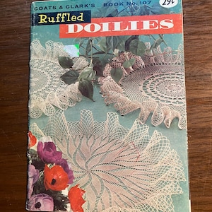 以下が含まれることがあります： ヴィンテージのクロシェドイリーのパターンブック、Coats & Clark's Book No. 107、タイトルは「Ruffled Doilies」、3つのドイリーと花の背景が特徴で、価格は29セントです。