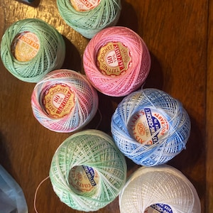 Puede incluir: Una colección de ovillos de hilo de tatting y ganchillo de colores en tonos verdes, rosas, azules y blancos. Cada ovillo tiene una etiqueta central con texto que indica el tipo de hilo, el metraje y la marca. Los hilos están bien enrollados, lo que sugiere que son nuevos.