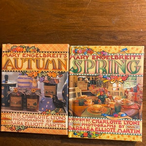 May include: Two hardcover books with colorful covers featuring illustrations of a kitchen scene. The book on the left is titled "Mary Engelbreit's Autumn" and the book on the right is titled "Mary Engelbreit's Spring". Both books are written by Charlotte Lyons and photographed by Barbara Elliott Martin.