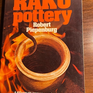 Op de afbeelding: Een boek getiteld "RAKU pottery" van Robert Piepenburg. De omslag toont een close-up van een keramische pot die in een oven wordt gebakken, omringd door vlammen. De tekst op de omslag luidt "Een 400 jaar oude Japanse keramische techniek aangepast voor de hedendaagse pottenbakker."