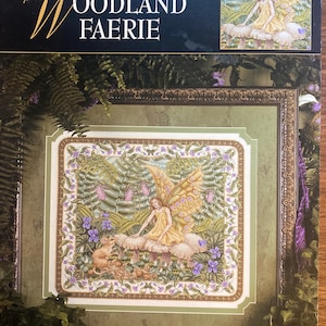 Könnte beinhalten: Ein Kreuzstich-Musterheft von Leisure Arts mit dem Titel Teresa Wentzler's Woodland Faerie. Das Cover zeigt ein fertiges Kreuzstichmotiv einer Fee mit gelben Flügeln, die auf Pilzen neben einer kleinen Maus sitzt, umgeben von Farnen und violetten Blumen.