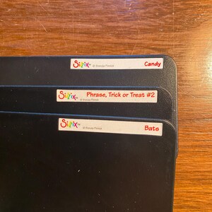 May include: Three black storage boxes with white labels. The labels have the text "Sizix" and the copyright symbol, followed by "Brenda Pinnick". The top label says "Candy", the middle label says "Phrase, Trick or Treat #2", and the bottom label says "Bats".