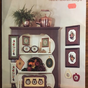 Op de afbeelding: Een vintage boekomslag voor een collectie kruissteekpatronen genaamd "Fruit Ambrosia" van Helene Halverson Designs. De omslag toont een koperen theepot, een varenplant en een houten plank met verschillende kruissteekontwerpen met fruit- en bloemenmotieven. De tekst "Leaflet 3 $4.00" is afgedrukt in de rechterbenedenhoek.