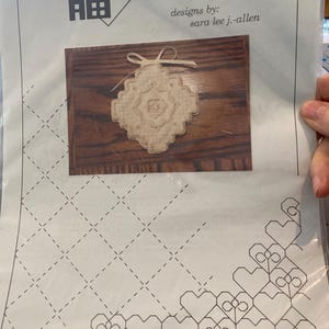 Op de afbeelding: Een knutselpakket getiteld "sew fun" met een kruissteekontwerp. De kit bevat een beige, geometrisch kruissteekstuk met een lint, tegen een houten achtergrond. De verpakking heeft een huis- en hartlogo en de naam van de ontwerper is Sara Lee J.-Allen.